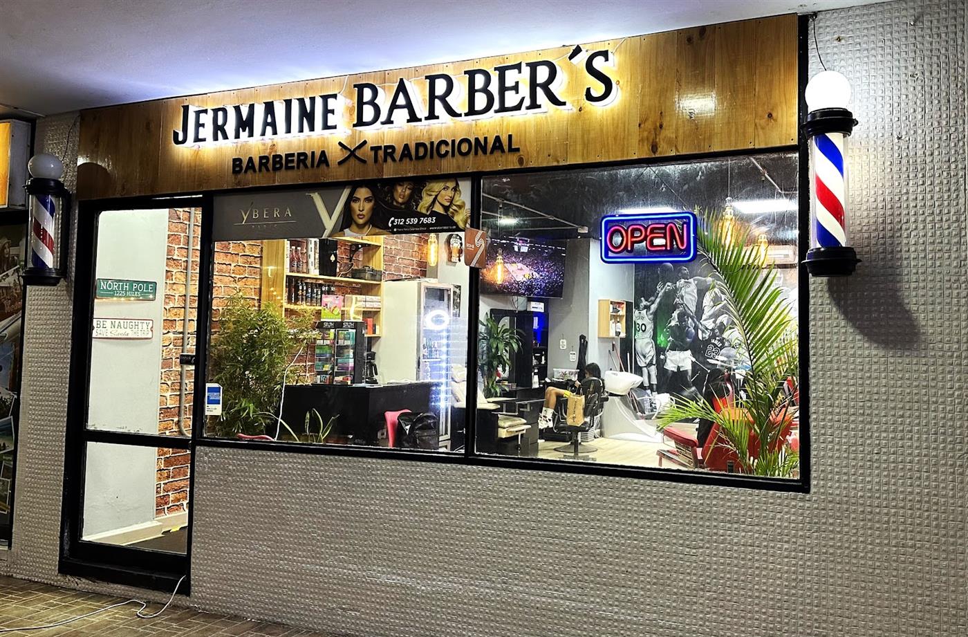 Fachada de Jermaine Barber's de noche, con el letrero de madera y el poste de barbero
